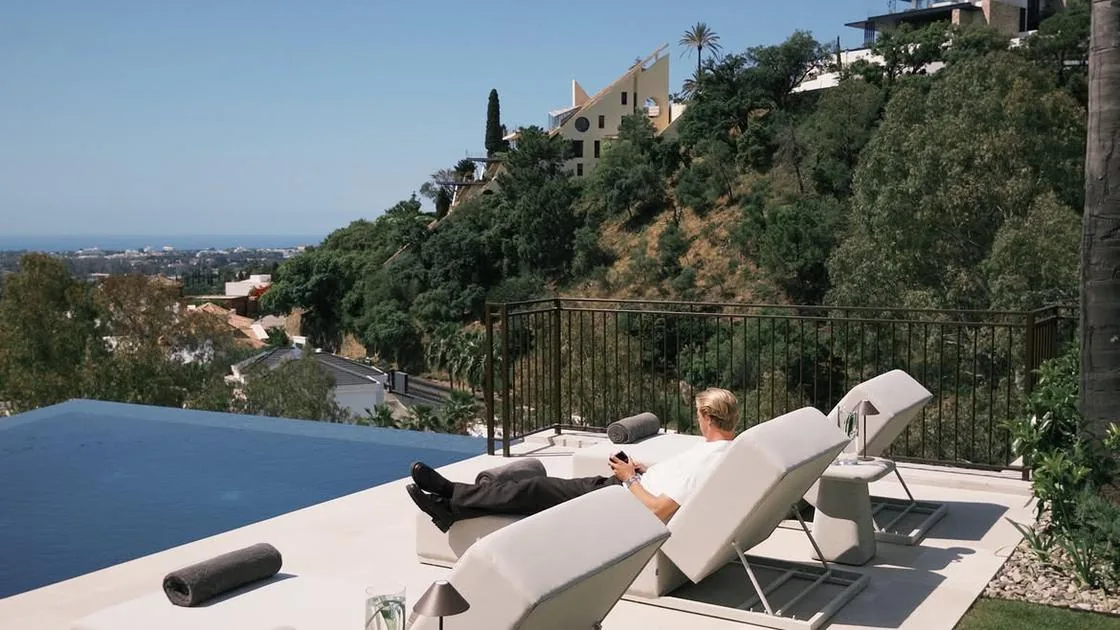 Rens Depraetere on a sun lounger beside a pool on a hillside terrace, phone in hand, with the sea in the distance